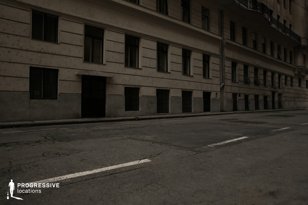 This narrow, worn-out street is lined with a plain, aged building featuring a repetitive window pattern and a neutral-toned façade. The lower-level windows are reinforced with metal bars, and the dark, uninviting entrance contributes to an atmosphere of neglect. The cracked asphalt, faded road markings, and lack of visible activity enhance the setting’s bleak and isolated feel. The location is well-suited for crime dramas, psychological thrillers, or dystopian films, offering a raw, unembellished urban environment ideal for scenes conveying tension, mystery, or urban decay.