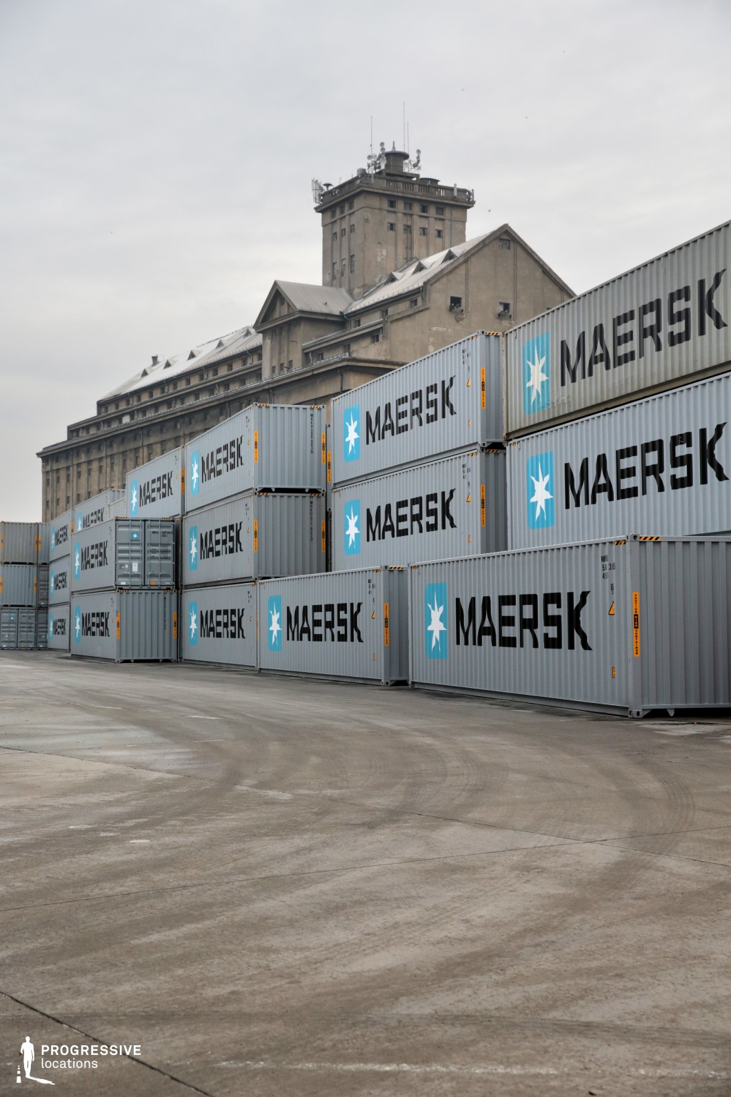 This image showcases a large industrial shipping yard with stacks of uniform shipping containers in muted gray tones, bearing bold branding. The rigid, repetitive geometry of the containers contrasts with the imposing historic warehouse in the background, which features a weathered concrete façade, a sloped roof with small dormer windows, and a prominent tower adorned with communication equipment. The open concrete ground in the foreground provides a sense of scale and emptiness, enhancing the stark industrial atmosphere. This location is well-suited for action thrillers, crime dramas, or espionage films requiring an isolated, industrial setting with a sense of secrecy and logistical activity.