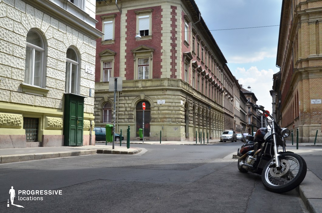 This European inner-city intersection is framed by grand historic buildings with a mix of textured stone and red-brick facades, featuring arched windows and decorative cornices. The architectural style blends late 19th-century and early 20th-century influences, creating a sense of urban depth and history. The narrow streets, flanked by green bollards and vintage-style street signs, add to the authentic old-world atmosphere. The location's enclosed layout and balanced symmetry make it ideal for crime dramas, period films, or urban thrillers, offering a cinematic backdrop rich in texture and historic character.