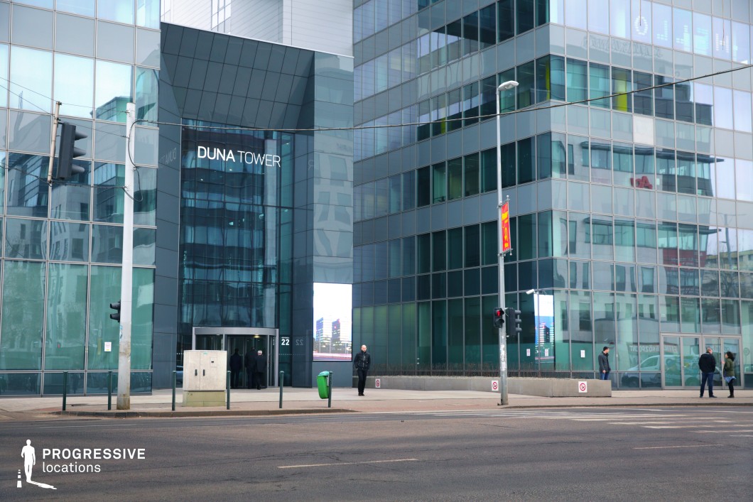 This modern high-rise office building features a sleek glass facade, reflecting the surrounding urban landscape and reinforcing a contemporary corporate aesthetic. The geometric design, clean lines, and tinted windows create an atmosphere of professionalism and exclusivity. The wide road in the foreground and minimalistic entrance plaza enhance the structured and business-oriented setting. This location is ideal for corporate dramas, financial thrillers, or espionage films, offering a polished and high-stakes backdrop for narratives centered around power, technology, or global business intrigue.