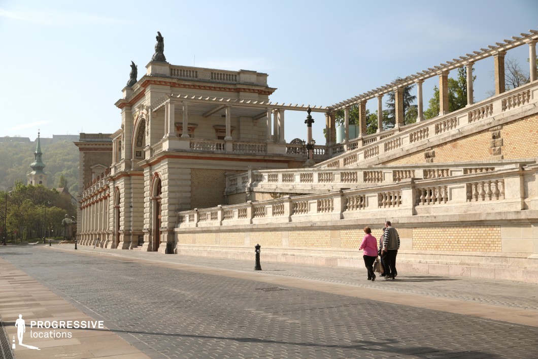 This image features an opulent neo-Renaissance architectural ensemble with a grand staircase, ornate balustrades, and decorative statues. The building’s intricate façade showcases detailed masonry, arched windows, and elegant columns, while warm-toned brick and stone create a harmonious blend of textures. A pergola-like structure extends along the upper terrace, adding a rhythmic repetition of classical elements. The sloping design of the stairway leads the eye upward, enhancing the sense of grandeur. In the background, a historic church spire rises above lush greenery, reinforcing the location’s historical depth. This setting is ideal for period dramas, aristocratic or historical narratives, and artistic productions requiring a refined European ambiance.