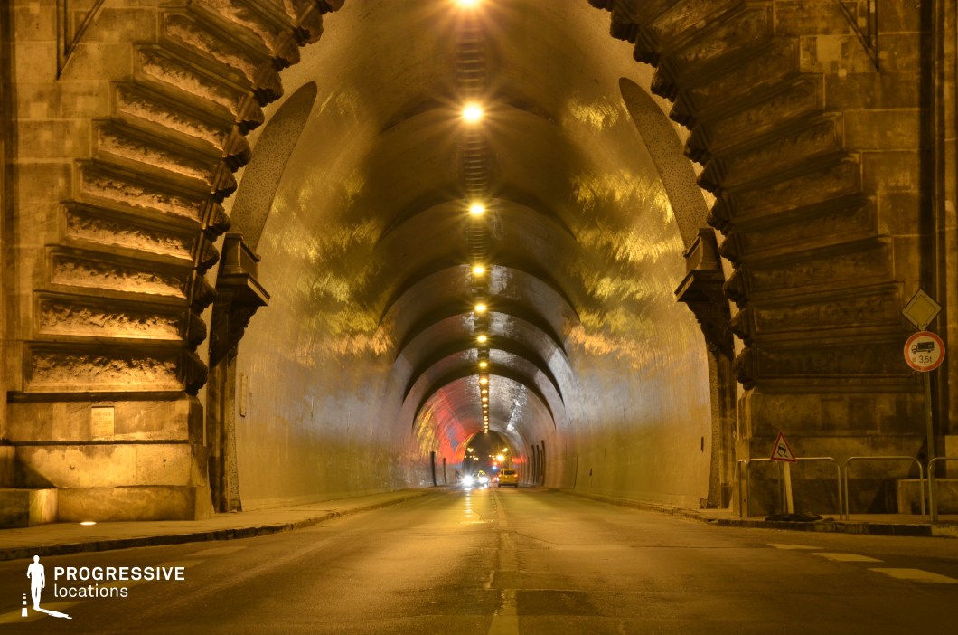 This grand, arched tunnel features intricately carved stone entrance details and a long, curving interior lined with warm, golden lighting. The tunnel’s reflective, smooth walls enhance the depth and repetition of its arched ceiling, creating a visually striking perspective. The soft glow from the overhead lamps contrasts with the dark road, adding an atmospheric intensity. The tunnel's historic stone construction and sense of enclosure make it an ideal setting for action sequences, car chases, or suspenseful thriller moments, offering a cinematic blend of grandeur and mystery.