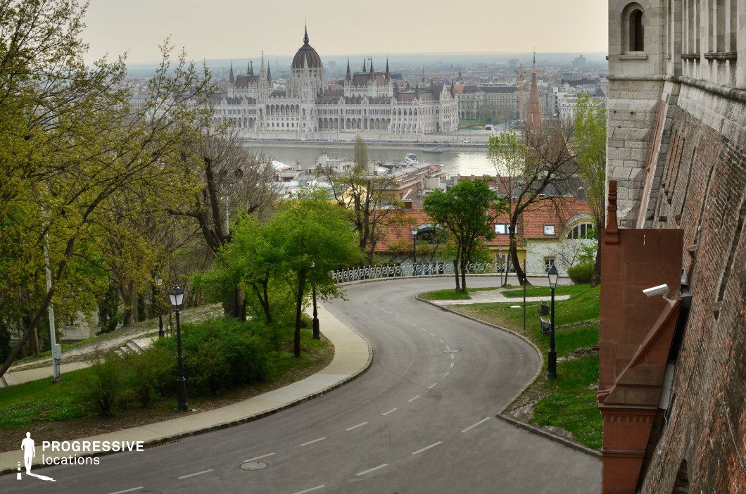 This elevated urban viewpoint captures a gracefully curving road framed by lush greenery and historic architecture, leading toward an expansive river and an iconic neo-Gothic building in the distance. The composition balances the natural elements of trees and park-like landscaping with the strong visual presence of stone and brick facades, creating a harmonious blend of urban and scenic aesthetics. The road’s gentle bend adds a dynamic perspective, making it an ideal setting for cinematic tracking shots, contemplative walking scenes, or sophisticated establishing shots for historical or dramatic films.