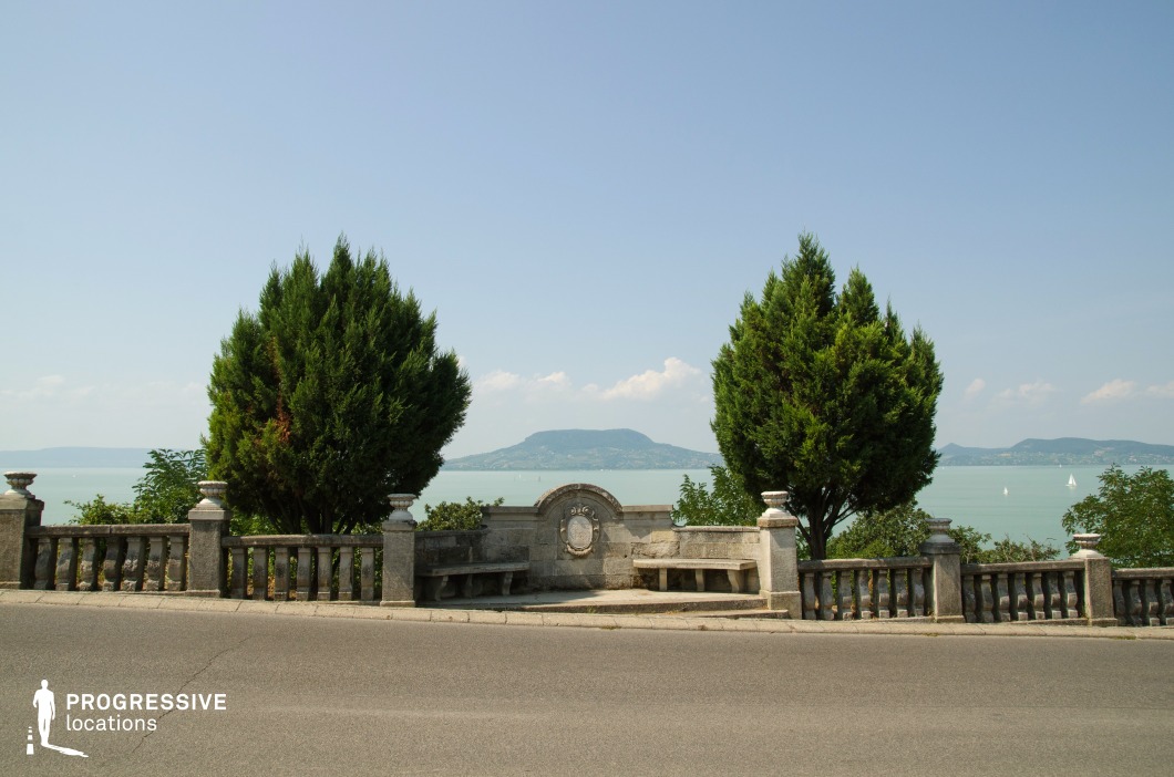 This image presents a picturesque vantage point overlooking a vast lake, framed by two symmetrically placed evergreen trees. The historic stone balustrade and benches add a classical elegance, while the central crest detail enhances the location’s refined character. In the background, a prominent volcanic hill rises above the water, contributing to the depth and natural grandeur of the setting. The scene exudes tranquility, making it an ideal location for romantic dramas, introspective moments in character-driven narratives, or establishing shots that emphasize serenity and scenic beauty.