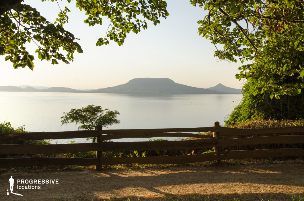 This image captures a tranquil lakeside viewpoint framed by lush green foliage and a rustic wooden fence. The still water extends toward the horizon, where a distinct flat-topped volcanic hill dominates the background, accompanied by smaller peaks and rolling hills. The foreground, with its dirt pathway and shaded vegetation, adds depth and natural framing to the composition. The setting exudes a peaceful, timeless atmosphere, making it an ideal location for contemplative or romantic scenes, nature documentaries, or establishing shots in films that emphasize serene landscapes and reflective moments.