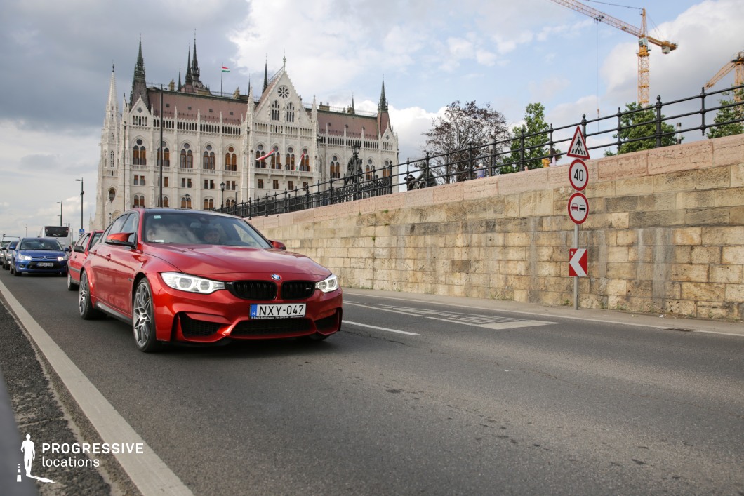 This dynamic city scene captures a well-trafficked roadway running parallel to a historic stone wall, beyond which stands an ornate neo-Gothic building with towering spires and intricate architectural details. The contrast between modern vehicles and the historic backdrop emphasizes the blending of contemporary urban life with classical architecture. The road’s perspective, marked by its dividing lines and structured barriers, creates a strong visual direction, making this location ideal for high-speed sequences, dramatic confrontations, or establishing shots in action, crime, or political thrillers.