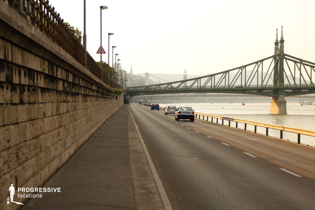 This long, curving riverside road runs parallel to the water, bordered by a textured stone retaining wall with ornate metal railings on one side and a protective barrier separating it from the river on the other. The dominating feature is the green metal truss bridge, with intricate spires and a sturdy industrial design, adding a strong visual anchor to the scene. In the background, the silhouettes of hills and historic buildings enhance the depth of the setting. The location is well-suited for dynamic filming sequences such as car chases, contemplative driving scenes, or establishing shots in a European city with a balance of historic and industrial elements.