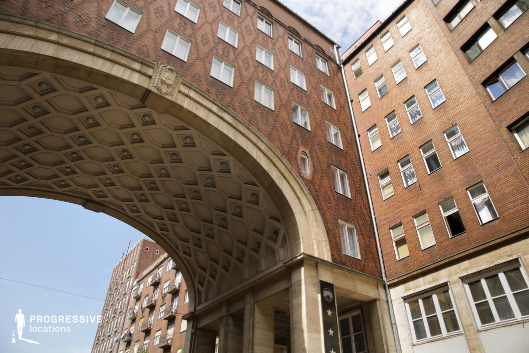 This monumental red-brick archway, adorned with an intricate diamond-patterned façade and coffered ceiling, creates a striking architectural focal point. The arch is framed by tall residential buildings with a uniform yet imposing appearance, featuring repetitive window arrangements that emphasize verticality. The blend of traditional brickwork and modernist influences lends the location a strong, institutional aesthetic, making it ideal for cinematic settings that require a sense of authority, urban resilience, or bureaucratic formality. This location suits political thrillers, dystopian narratives, or historical films depicting mid-century European cityscapes.