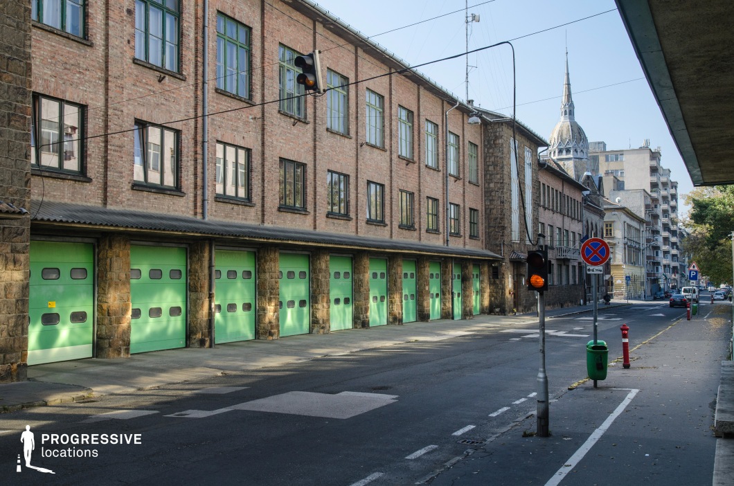 This image captures a long, industrial-style brick building featuring a row of large green garage doors, framed by stone columns that add a rugged, utilitarian aesthetic. The upper level showcases a series of rectangular windows with dark green trim, reinforcing the structured, repetitive design. Overhead, traffic lights and street signs contribute to the urban atmosphere, while a distant Gothic-style spire rises above the surrounding structures, adding an unexpected architectural contrast. The slightly worn street and sidewalk enhance the industrial character of the setting. This location is well-suited for crime dramas, investigative thrillers, or action sequences, particularly those involving covert operations or chase scenes through an urban industrial backdrop.