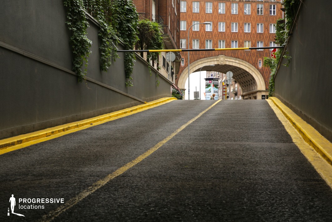 This upward-sloping urban driveway, flanked by ivy-covered walls, leads toward a grand arched passage that frames the city beyond. The textured asphalt, with its bold yellow dividing lines and curbstones, adds a sense of depth and direction. The surrounding architecture combines modern and historic elements, with a patterned brick façade and ornate stone arch serving as a striking visual contrast. The tight, enclosed nature of the ramp, coupled with the dramatic rise toward an open street, makes this location ideal for suspenseful chase scenes, psychological thrillers, or moody urban dramas requiring a visually compelling transition space.