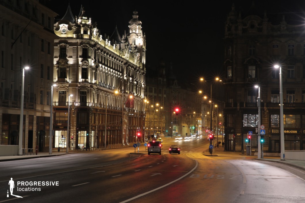 This sweeping European boulevard at night features a striking contrast between ornate, illuminated historic buildings and a wide, modern roadway. The intricate façade of the corner building, with its decorative turrets and classical detailing, stands out as a focal point against the dark night sky. The gentle curve of the road, lined with evenly spaced streetlights and traffic signals, creates a cinematic depth, while reflections on the wet pavement add a layer of atmospheric elegance. This location is ideal for high-end drama, noir-style thrillers, or luxury brand commercials, offering a sophisticated urban setting with a sense of grandeur and timelessness.