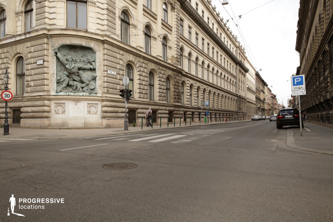 This wide European boulevard is lined with imposing neoclassical buildings featuring decorative stonework, arched windows, and intricate cornices. A striking bronze relief sculpture embedded in the corner façade adds historical significance and artistic depth to the scene. The long, straight road is framed by evenly spaced bollards and tram cables overhead, reinforcing the city's structured and historic character. The balanced composition, with a mix of old-world charm and functional urban infrastructure, makes this location ideal for period dramas, political thrillers, or establishing shots of an elegant European setting with a sense of authority and history.
