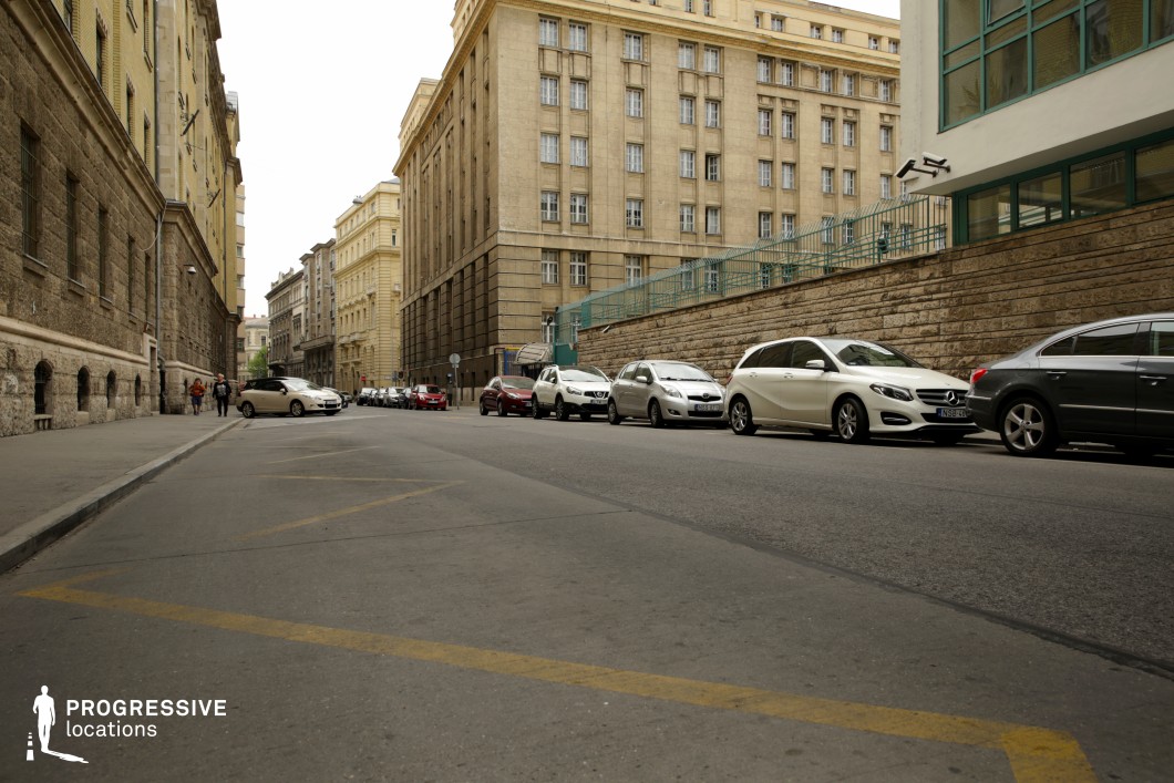 This urban street is flanked by imposing, historic buildings featuring a mix of neoclassical and early 20th-century institutional architectural styles. The facades are constructed from stone and plaster, with a subdued color palette of beige, brown, and muted yellow, contributing to a formal, almost austere atmosphere. The street itself is relatively wide, with clear road markings and a gently curving sidewalk, providing a sense of depth and perspective. A stone retaining wall with a metal railing on one side adds a structured, fortified feel, while the uniform rows of tall windows emphasize the verticality of the buildings. The location is ideal for film productions requiring a European institutional or governmental setting, suitable for political thrillers, espionage dramas, or historical reconstructions.
