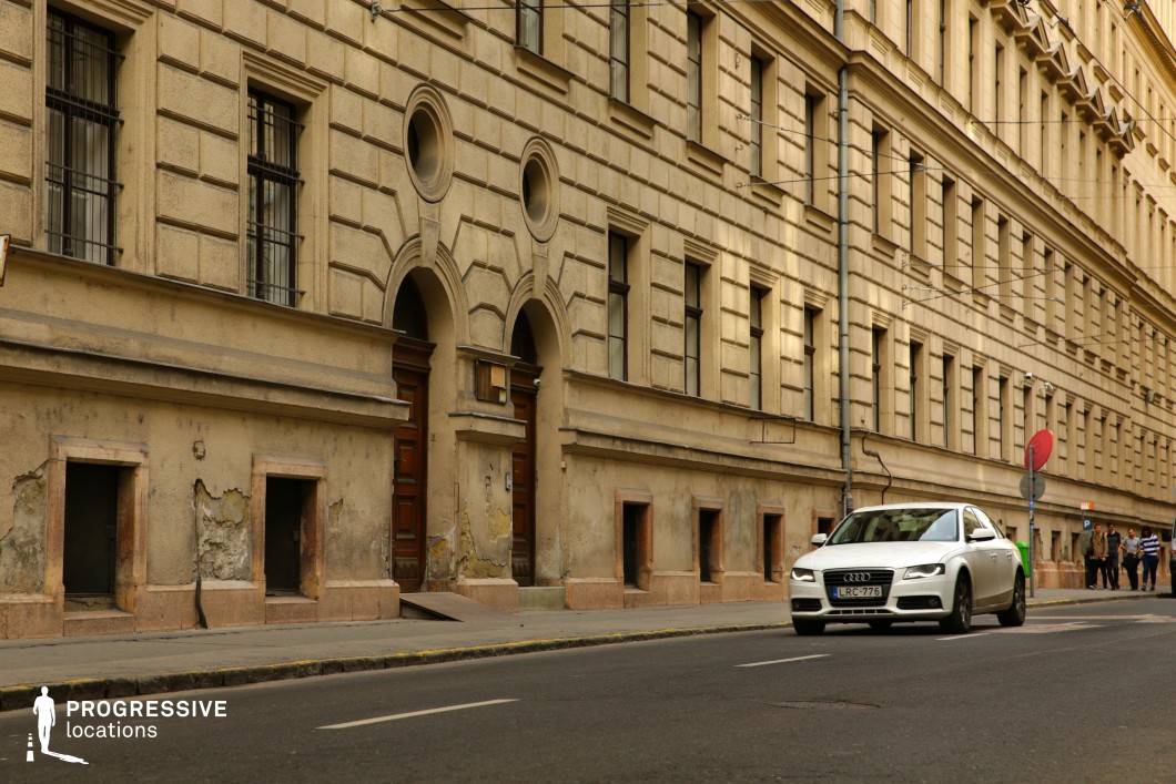 This urban street is lined with an aged neoclassical-style building featuring a textured façade with intricate geometric patterns, arched doorways, and small, barred windows. The weathered plaster and exposed brickwork add a sense of character and history to the structure. The street’s perspective leads into the distance, flanked by modern vehicles and pedestrians, creating a natural sense of depth. This setting is ideal for historical dramas, noir-style thrillers, or urban storytelling that requires an atmospheric, lived-in European backdrop with an air of mystery and nostalgia.