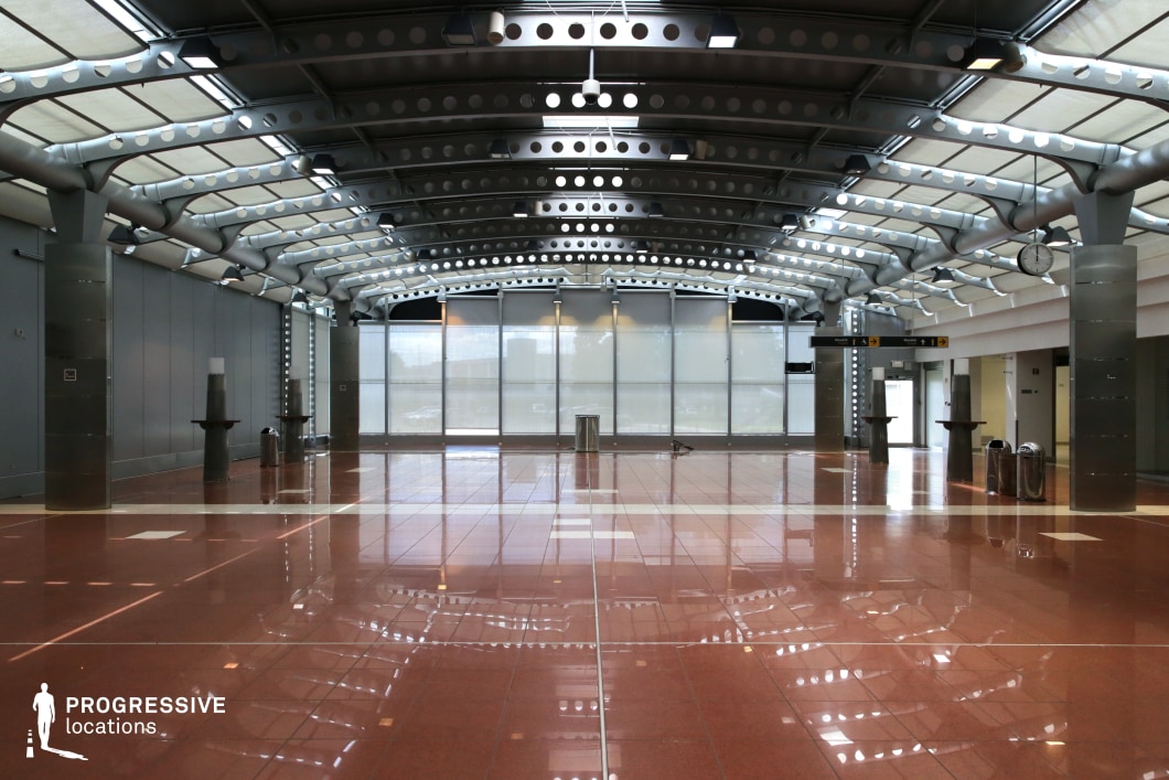This sleek, futuristic terminal space is defined by its industrial aesthetic, featuring a curved metal ceiling with perforated steel beams and exposed structural supports. The polished reddish-brown tiled floor reflects the overhead lighting, adding depth to the space. Large frosted glass windows diffuse natural light, creating a clean, modern ambiance. Minimalist metal columns and directional signage reinforce the location’s functional design, reminiscent of an airport or transit hub. This setting is ideal for sci-fi, action, or espionage thrillers, providing a high-tech, corporate, or security-heavy atmosphere, perfect for tense meetings, chases, or high-stakes confrontations.