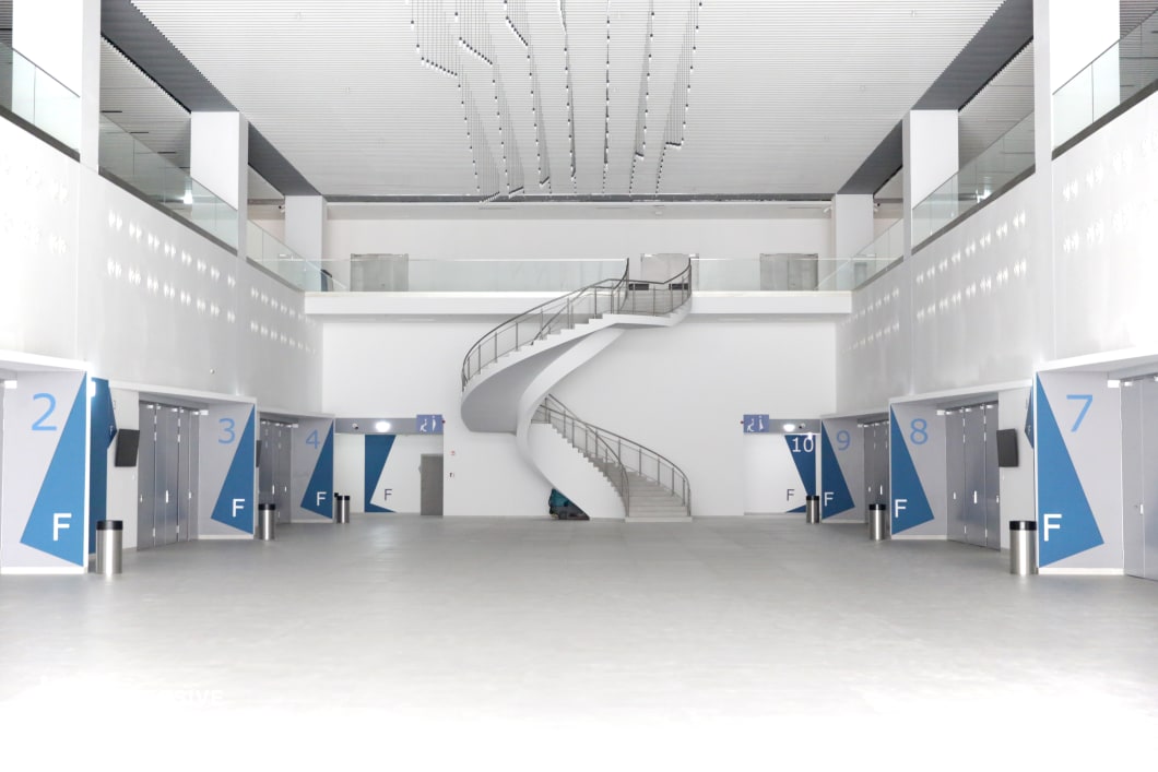 This expansive, high-ceilinged terminal features a clean, futuristic design dominated by stark white walls and geometric blue signage marking numbered gates. A sculptural spiral staircase at the center serves as a striking focal point, leading to an upper mezzanine enclosed by glass railings. The polished light-gray flooring and sleek ceiling lighting contribute to the sterile, ultra-modern aesthetic. The symmetrical layout, open space, and minimalist architecture create an atmosphere that is both functional and cinematic. This location is ideal for sci-fi productions, corporate thrillers, or high-tech espionage films, offering a versatile backdrop for transit-related scenes, security checkpoints, or tense confrontations in a controlled, sterile environment.