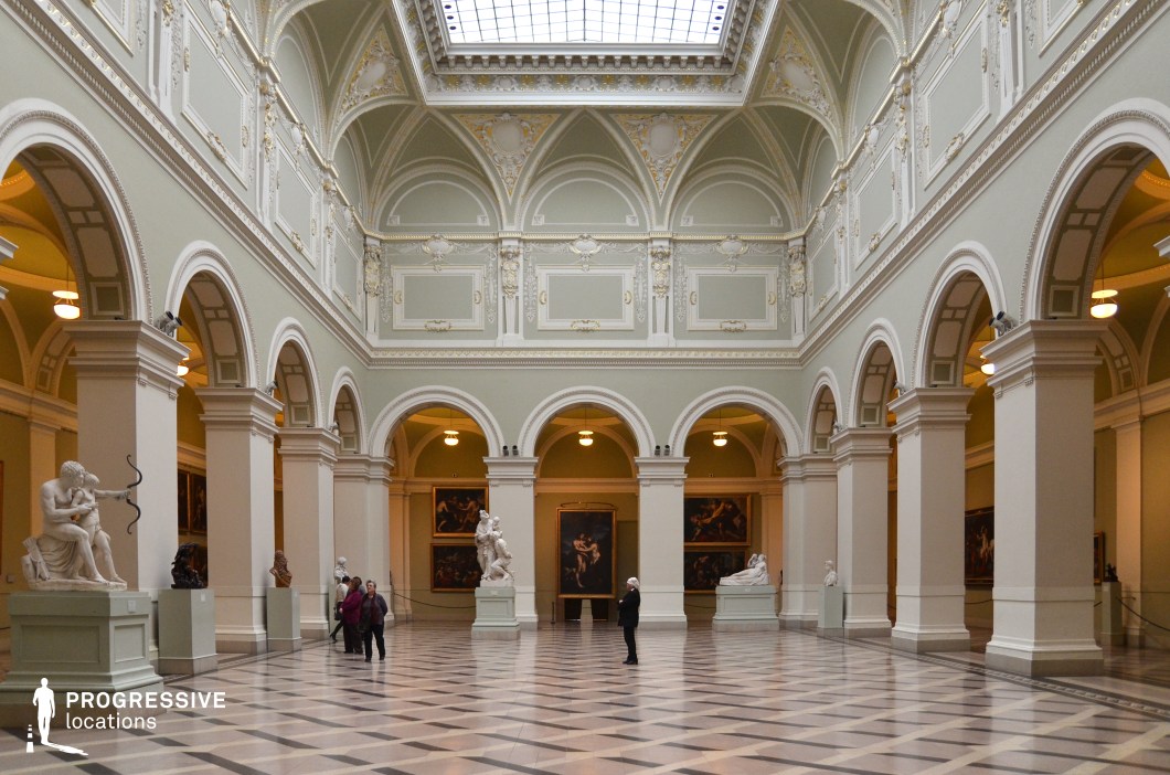 This grand museum hall showcases a refined neoclassical architectural style with towering arched openings, ornate moldings, and intricate gold-accented detailing. The high, vaulted ceiling is adorned with decorative panels, and a central skylight allows natural light to illuminate the space. The polished checkered marble floor enhances the hall's stately atmosphere, reflecting the surrounding columns and classical sculptures displayed on pedestals. The soft pastel hues of the walls contrast elegantly with the darker framed paintings in the adjoining galleries. This location is ideal for historical dramas, art-themed narratives, and sophisticated intrigue, providing a regal and timeless setting for moments of reflection, discovery, or high-society gatherings.