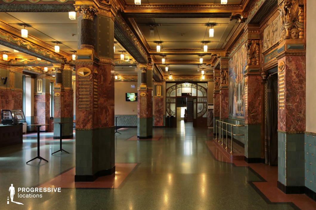 This grand Art Nouveau hall is characterized by its intricate architectural detailing, featuring elaborately decorated columns adorned with gilded reliefs, deep red marble, and green ceramic tiling. The ceiling boasts ornamental beams with gold-accented geometric patterns, complemented by hanging square-shaped light fixtures. Large murals depicting allegorical scenes enhance the walls, framed by sculpted faces and mythical griffins that add to the hall’s regal atmosphere. At the far end, a striking arched doorway with frosted glass panels and wrought ironwork leads to an upper-level staircase, enhancing the sense of depth. This location is ideal for filming period dramas, historical epics, or fantasy productions, providing a lavish and immersive setting for high-society gatherings, political intrigue, or mysterious encounters.