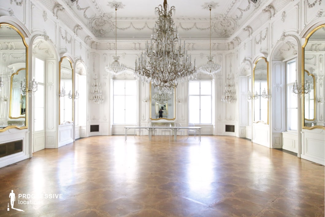 This grand ballroom features an opulent Rococo-inspired design with a predominantly white and gold color scheme, creating a luminous and sophisticated ambiance. High ceilings adorned with intricate stucco moldings complement the large arched mirrors framed in gold, enhancing the sense of space and symmetry. A striking crystal chandelier, along with several smaller chandeliers, dominates the center, reflecting light across the polished parquet floor. Large windows allow for natural illumination, further amplifying the room’s ethereal quality. This location is ideal for filming scenes requiring a regal or aristocratic setting, making it perfect for period dramas, high-society gatherings, or romantic ballroom sequences.