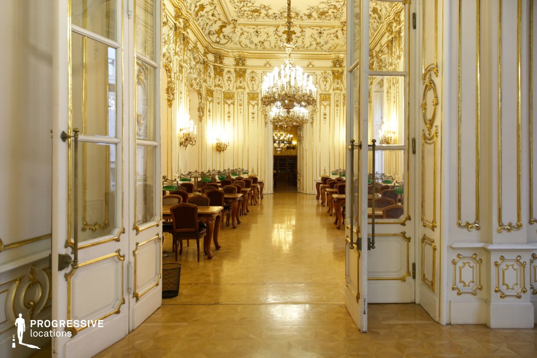 This grand hall showcases an opulent Baroque interior, featuring gilded moldings, elaborate stucco details, and multiple chandeliers that illuminate the space with a warm glow. The high, decorated ceiling enhances the sense of grandeur, while the symmetrical layout of wooden tables and elegant chairs suggests a refined library or gathering space. Ornate wall sconces line the room, adding depth and sophistication. The polished parquet flooring reflects the intricate architectural details, further amplifying the room’s elegance. This location is ideal for filming historical dramas, aristocratic social gatherings, or period-piece intellectual debates, offering a setting rich in prestige and old-world charm.