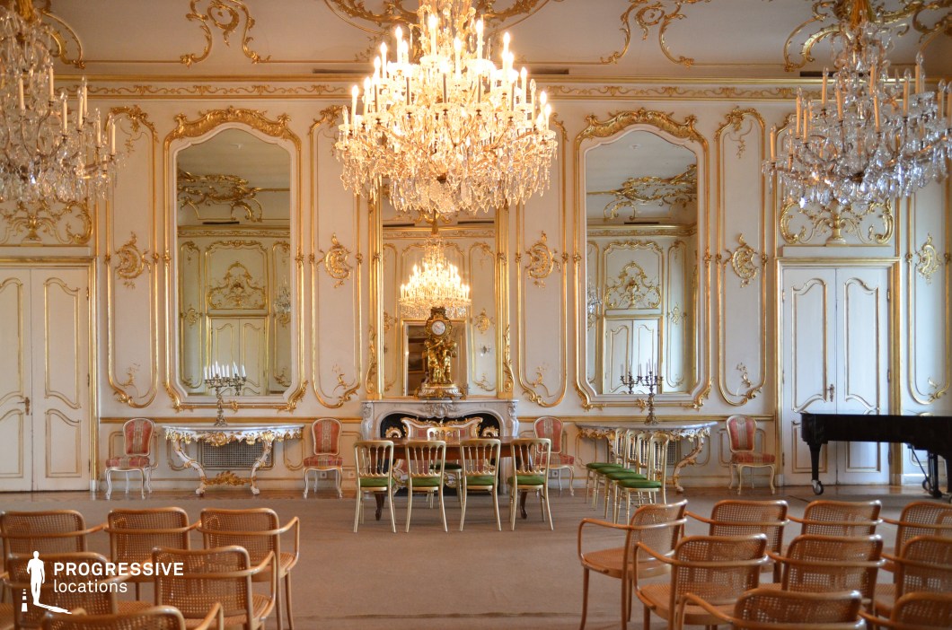 This grand Baroque-style ballroom exudes opulence with its intricate gold-leaf moldings, tall mirrors framed in gilded detailing, and multiple crystal chandeliers casting a warm glow. The symmetrical design is accentuated by a central fireplace with an ornate clock, flanked by luxurious seating and elaborately carved furniture. The high ceilings and cream-colored walls create an airy yet regal ambiance. A black grand piano sits in the corner, enhancing the room’s function as a performance or ceremonial space. The setting is ideal for filming historical dramas, aristocratic gatherings, period-piece ballroom scenes, or luxury brand promotions, offering an extravagant and refined aesthetic.