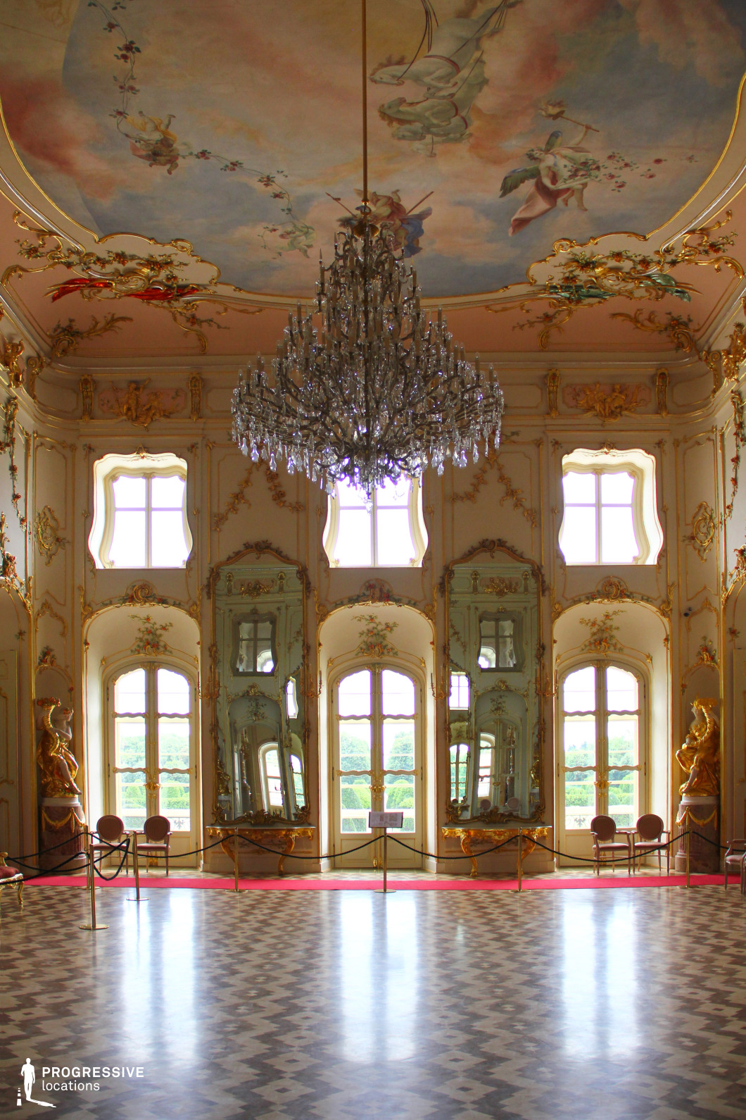 This extravagant Rococo-style ballroom features soaring ceilings adorned with hand-painted frescoes of cherubs and allegorical figures, framed by intricate gold leaf moldings. A grand crystal chandelier hangs at the center, reflecting light across the space. The tall arched windows and mirrors, decorated with gilded details and pastel accents, amplify the sense of grandeur and create an illusion of expanded depth. The checkered marble floor contrasts elegantly with the ornate surroundings, while statues and delicate wall embellishments reinforce the aristocratic ambiance. This setting is perfect for filming period dramas, historical romances, royal court scenes, and luxury fashion productions, offering a rich, visually immersive backdrop.