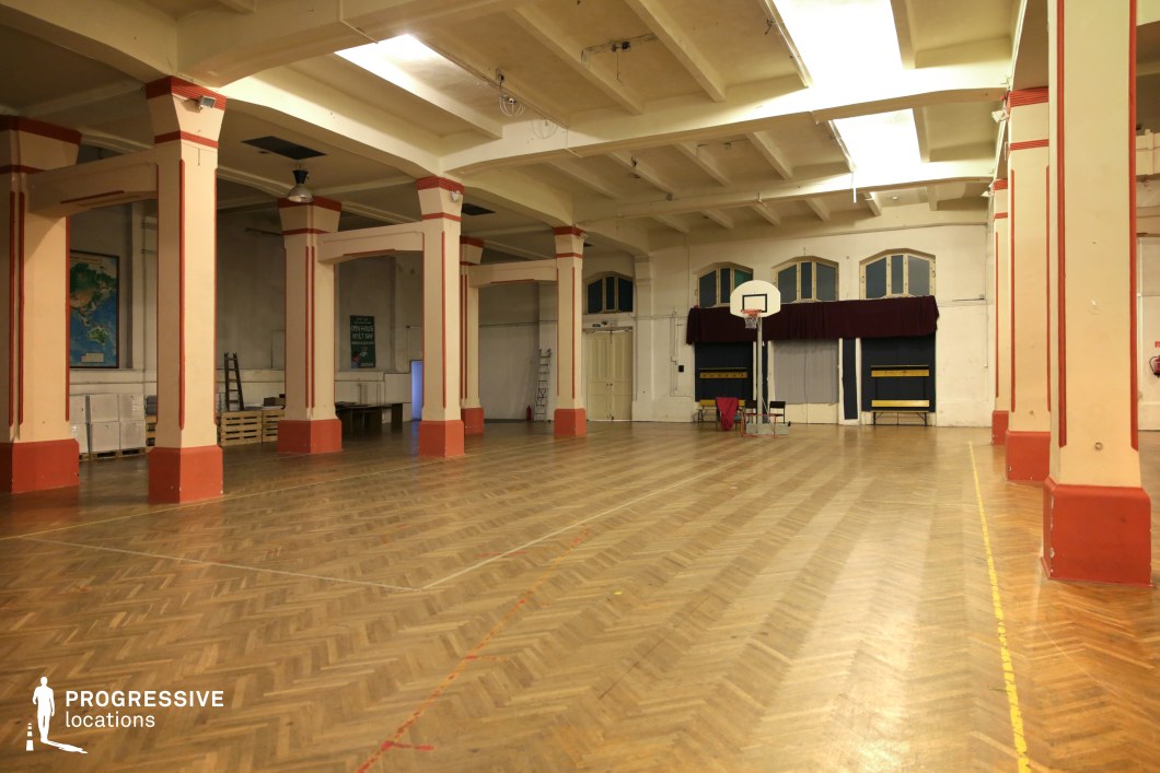 This spacious indoor gymnasium features a retro-industrial design with thick structural columns accented in red, a high coffered ceiling, and a classic herringbone-patterned wooden floor marked for sports activities. A single basketball hoop stands against an arched-windowed wall, adding to the aged institutional atmosphere. The worn yet functional aesthetic, along with old maps and wooden pallets along the walls, suggests a repurposed or long-standing community space. The large open floor area and strong architectural symmetry make this location ideal for filming nostalgic school settings, underground sports scenes, or tense dramatic sequences in a raw, utilitarian environment.