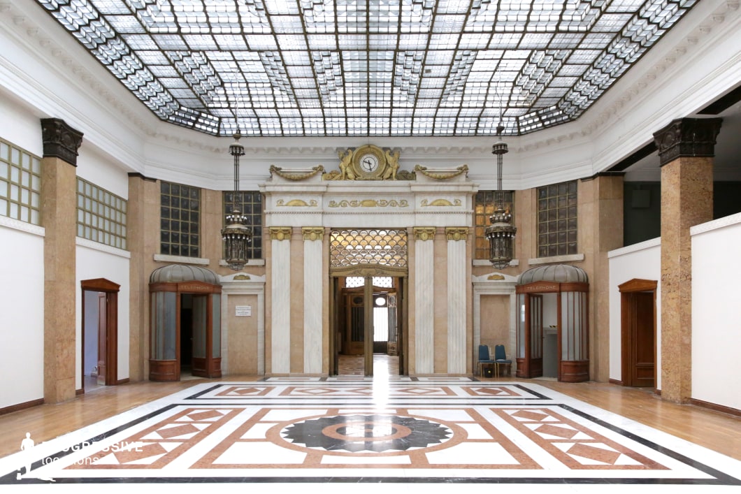 This opulent Art Deco hall features a striking glass ceiling that bathes the space in diffused natural light, highlighting the geometric patterns of the polished marble floor. The walls are adorned with grand marble columns, gold accents, and frosted glass panels, enhancing the luxurious aesthetic. The focal point is an ornate entrance framed by white and gold pilasters, topped with a sculptural relief and a decorative clock. Hanging lantern-style chandeliers contribute to the hall’s vintage grandeur, while classic wooden telephone booths add a nostalgic element. This location is ideal for filming historical dramas, noir thrillers, or sophisticated period pieces, offering a refined setting for power dynamics, high-stakes meetings, or mystery-driven narratives.