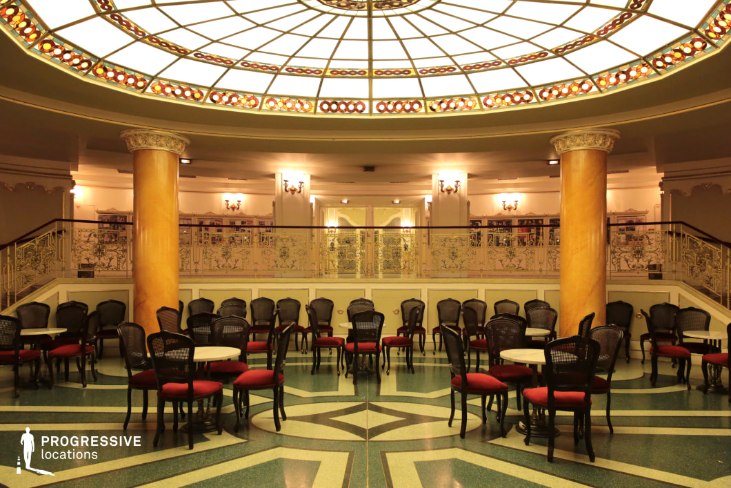 This opulent lounge showcases classic European elegance, featuring a grand oval stained-glass ceiling that diffuses warm ambient light. The space is defined by two large, polished columns and intricate wrought-iron railings on the upper level, adding to its refined aesthetic. The richly patterned terrazzo flooring, paired with dark wooden chairs upholstered in deep red fabric, creates a luxurious yet inviting atmosphere. Ornate wall sconces provide additional illumination, enhancing the classical décor. This location is ideal for filming historical dramas, period romances, or high-society intrigue, as well as casino, ballroom, or grand café scenes set in the early to mid-20th century.