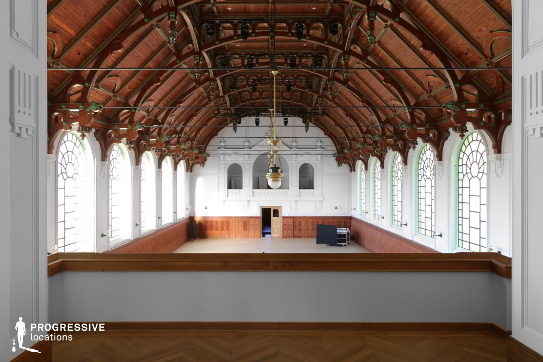 This striking hall features an expansive wooden vaulted ceiling supported by intricate green and bronze metal brackets, lending a blend of industrial elegance and classical refinement. The high arched windows, framed by white stucco, allow natural light to flood the space, accentuating the rich wood paneling along the lower walls. The symmetrical design leads the eye toward an ornate white balcony with sculptural detailing above the central entrance. A grand brass pendant light hangs from the ceiling, reinforcing the historic character of the hall. This location is ideal for filming historical dramas, classical performances, or formal events, offering an elegant yet spacious setting suitable for grand ceremonies, aristocratic gatherings, or period-piece storytelling.