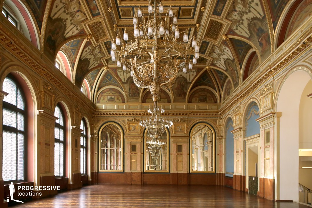 This grand ballroom embodies classical opulence, featuring elaborately painted frescoes on its arched ceilings, intricate gold detailing, and a series of grand chandeliers that add a regal ambiance. The large arched windows, framed by elegant moldings, allow natural light to flood the space, enhancing the warm tones of the parquet flooring and the intricate patterns on the walls. Tall mirrors, set within gilded frames, create a sense of depth and continuity, reflecting the room’s grandeur. The combination of rich architectural details and refined symmetry makes this location perfect for filming historical dramas, period romance films, royal ceremonies, or high-society gatherings, providing a sophisticated and visually stunning backdrop.