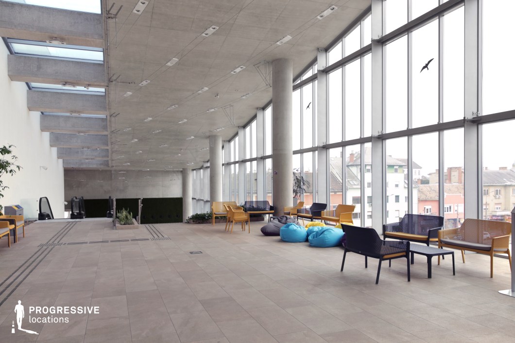 This modern, industrial-style lounge features an open-concept design with floor-to-ceiling windows that offer expansive views of the surrounding urban landscape. The raw concrete ceiling, exposed beams, and structural pillars create a minimalist, almost brutalist aesthetic, softened by warm wooden seating and colorful bean bags. The neutral-toned tiled flooring adds to the clean and spacious atmosphere, while greenery introduces a subtle natural element. The large glass facade allows for abundant natural light, making this an ideal location for contemporary urban scenes, corporate dramas, or futuristic and dystopian narratives. The setting is versatile for scenes involving business meetings, casual interactions, or high-tech environments.