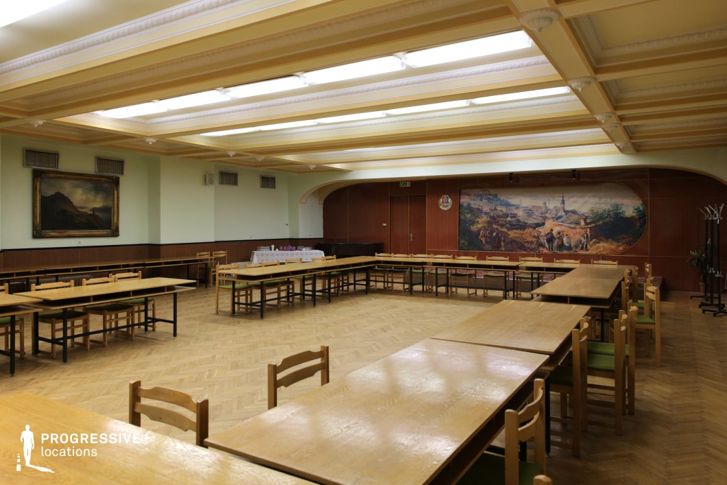 This spacious meeting hall features a mid-20th-century institutional aesthetic, with a structured coffered ceiling housing fluorescent lighting. The room is furnished with long wooden tables and simple wooden chairs with green upholstered seats, arranged in a flexible, communal layout. The parquet flooring adds warmth, while the walls are adorned with wood paneling and pastel-colored sections. A large mural depicting a historic rural scene serves as a focal point, reinforcing a traditional or civic atmosphere. The setting suggests a formal yet modest gathering space, making it ideal for filming political or historical dramas, community meetings, or retro institutional scenes set in an Eastern European or Soviet-era context.