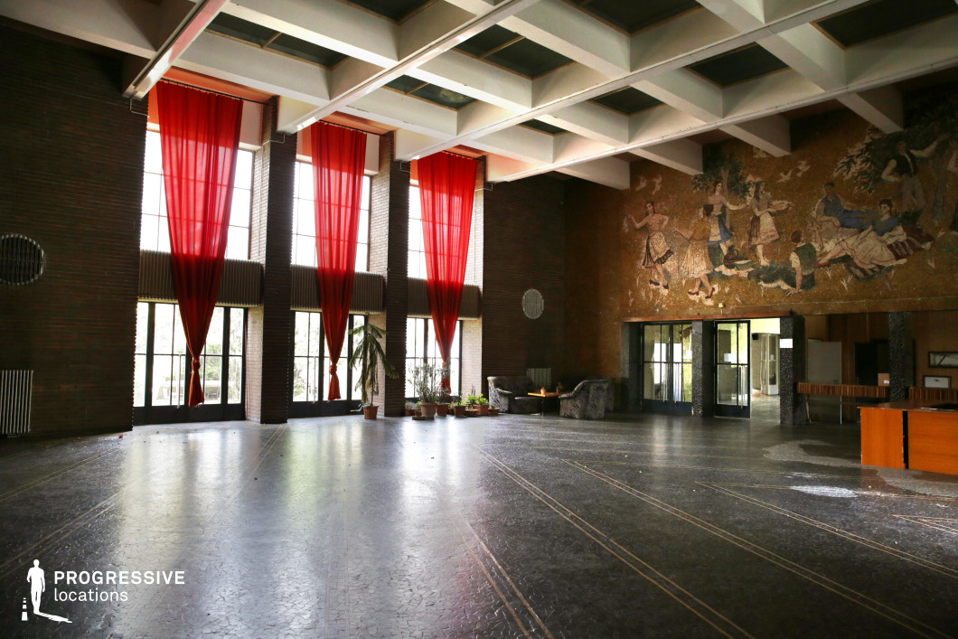 This grand cultural hall features a spacious open floor plan, accentuated by tall floor-to-ceiling windows draped in striking red curtains that contrast against the exposed brick walls. The high ceiling incorporates a geometric grid pattern, lending a structured yet airy feel to the space. A large mosaic mural on one wall depicts an idealized pastoral scene, adding historical and artistic depth. The dark stone flooring is intersected with fine metallic inlays, enhancing the hall's vintage character. This location is well-suited for period films, political dramas, and historical documentaries, particularly those set in mid-20th-century Eastern Europe. It provides an ideal backdrop for scenes involving cultural events, formal receptions, or reflective moments in a grand yet slightly faded setting.