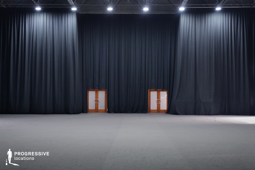This vast, open space features a black-box theater or studio setup, defined by towering black curtains that extend from the ceiling to the floor. The industrial ceiling structure and evenly spaced overhead lights create a controlled and adaptable lighting environment. Two wooden double doors with frosted glass panels contrast sharply against the dark backdrop, adding a subtle architectural element. The neutral flooring allows for full creative flexibility, making the space an ideal blank canvas for set design, theatrical performances, film productions, or experimental visuals. This location is perfect for avant-garde filmmaking, minimalist stage productions, psychological thrillers, and conceptual art pieces that require a stark, immersive setting.