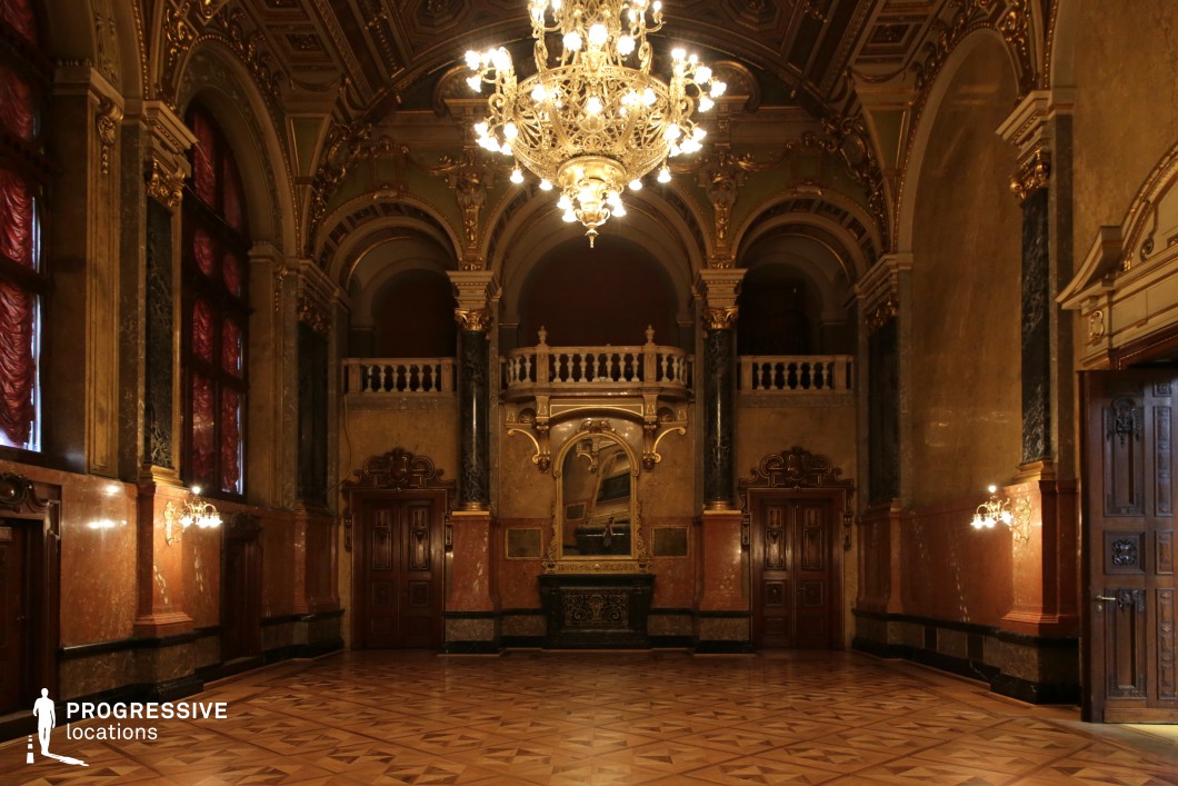 This opulent hall is defined by its rich Baroque detailing, dark marble columns, and warm golden accents. A grand chandelier dominates the space, casting a dramatic glow over the intricate parquet flooring and gilded wall sconces. The tall, arched windows are draped in deep red fabric, adding a regal touch to the room’s already imposing presence. Ornate balconies with carved stone balustrades overlook the main floor, enhancing the verticality of the architecture. The symmetrical layout and lavish decor make this location ideal for filming historical dramas, aristocratic gatherings, or scenes requiring a sense of mystery, grandeur, and old-world elegance.
