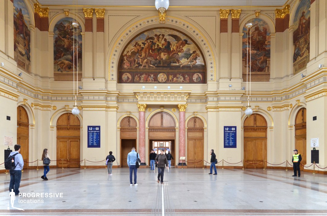 This grand hall showcases a richly decorated neoclassical interior, featuring soaring ceilings adorned with intricate frescoes, gold-accented moldings, and arched wooden doorways. Two striking pink marble columns frame the central entrance, while large semi-circular murals depict allegorical and historical scenes, adding depth and grandeur. The polished stone floor enhances the sense of space and elegance, while hanging globe lights provide soft illumination. The fusion of classical and baroque elements creates a setting ideal for historical dramas, period films, and formal sequences, offering a regal and culturally rich atmosphere suited for scenes of grandeur, diplomacy, or artistic appreciation.