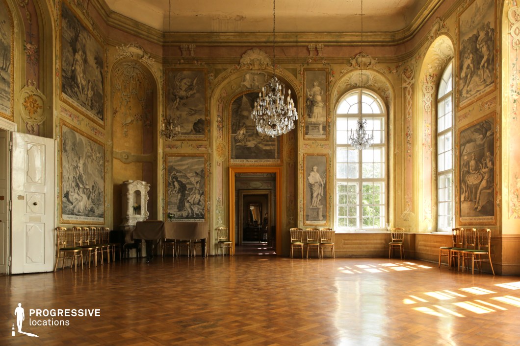 This grand ballroom exudes an aged elegance, featuring intricate frescoes depicting classical scenes framed by ornate floral moldings. The high, arched windows allow natural light to cast warm highlights on the parquet flooring, enhancing the room's depth and texture. Subtle pastel hues blend with faded gold accents, creating a timeless atmosphere. A central chandelier, along with additional smaller ones, adds a touch of refinement, while rows of antique chairs line the walls, emphasizing the space's historical significance. The long, open doorway leading into additional chambers enhances the sense of depth and continuity. This location is ideal for filming historical dramas, period romances, or artistic biopics, perfectly suited for scenes of aristocratic gatherings, secretive meetings, or grand but decayed elegance.