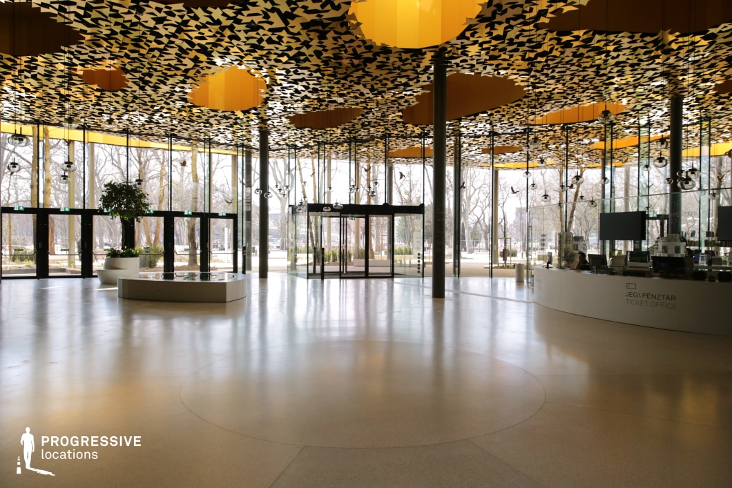 This spacious, modern pavilion features floor-to-ceiling glass walls that create an open and airy atmosphere, seamlessly blending the interior with the surrounding park landscape. The standout feature is the striking ceiling, adorned with an intricate golden geometric pattern that casts dynamic shadows across the polished light-colored floor. Slim, dark support columns enhance the sleek, minimalist aesthetic, while strategically placed hanging lights add to the ambiance. A curved ticket counter and planter installations introduce subtle architectural elements, maintaining a clean and futuristic design. This location is ideal for high-end commercial shoots, corporate settings, or futuristic and sci-fi productions, offering a visually distinctive space for sophisticated and concept-driven storytelling.