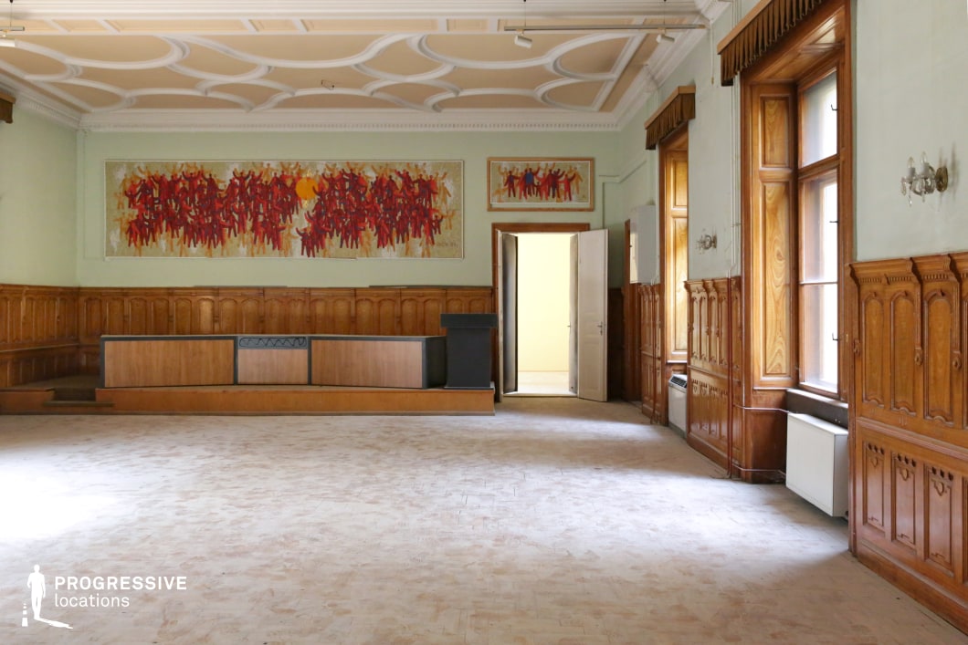 This spacious hall exudes a vintage institutional atmosphere with its high ceilings, decorative coffered plasterwork, and extensive wooden paneling that lines the walls. Large windows with deep wooden casings allow natural light to filter in, complementing the warm tones of the woodwork. The focal point is a raised platform at the far end, likely for speeches or presentations, framed by two bold, red-toned mosaic artworks that contrast with the subdued surroundings. The worn flooring and slightly aged elements add to the room’s historical charm. This location is ideal for filming period dramas, political or academic settings, or scenes requiring a grand yet slightly aged interior, fitting for historical reconstructions, suspenseful meetings, or intellectual debates.