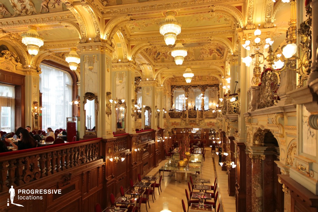This breathtaking café showcases an opulent Baroque-style interior, featuring soaring ceilings adorned with intricate gold detailing, sculpted moldings, and grand chandeliers that illuminate the space with a warm, elegant glow. The architectural symmetry is emphasized by polished marble columns, gilded balustrades, and decorative mirrors framed in gold, enhancing the depth and refinement of the setting. The upper-level balconies, adorned with statues and elaborate ironwork, overlook the main dining area, which is furnished with glass-topped tables and plush red seating, blending historical grandeur with modern sophistication. This location is ideal for filming high-society gatherings, period dramas, lavish romance films, or luxury lifestyle narratives, offering an extravagant and visually mesmerizing backdrop.