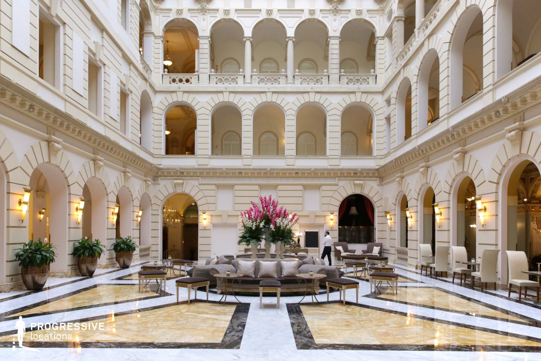 This refined courtyard blends neoclassical architecture with modern luxury, featuring a symmetrical design of grand arches, intricate moldings, and a soft cream-colored façade. The polished marble flooring showcases an eye-catching geometric pattern in gold, white, and black, enhancing the grandeur of the space. Elegant lounge seating and carefully arranged floral decorations add a sophisticated touch, while warm wall sconces create a welcoming ambiance. The open, multi-level structure, with ornate balconies and colonnades, evokes a sense of exclusivity and prestige. This location is ideal for filming high-society gatherings, luxury hotel sequences, or dramatic encounters in a setting of wealth and elegance, making it well-suited for romance, period dramas, or corporate thrillers.