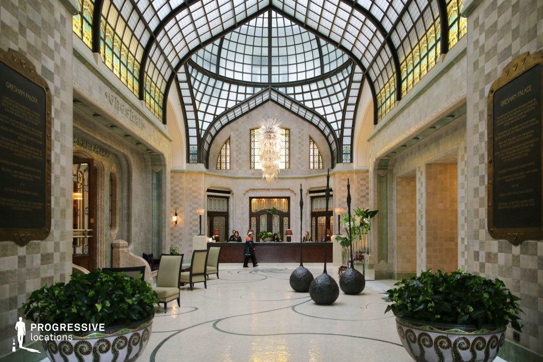 This refined Art Nouveau lobby features a striking glass-paneled ceiling with sweeping curves and intricate stained-glass accents that cast a soft, ambient glow. The symmetrical design, with grand arched windows and polished stone surfaces in neutral tones, creates an airy and sophisticated atmosphere. A contemporary glass chandelier serves as a central focal point, complementing the artistic mosaic flooring with delicate swirling patterns. Ornamental details, including sculptural decor and carefully arranged greenery, enhance the space’s refined aesthetic. This location is ideal for filming high-end luxury settings, period dramas, or stylish modern narratives, offering a timeless blend of architectural elegance and contemporary sophistication.
