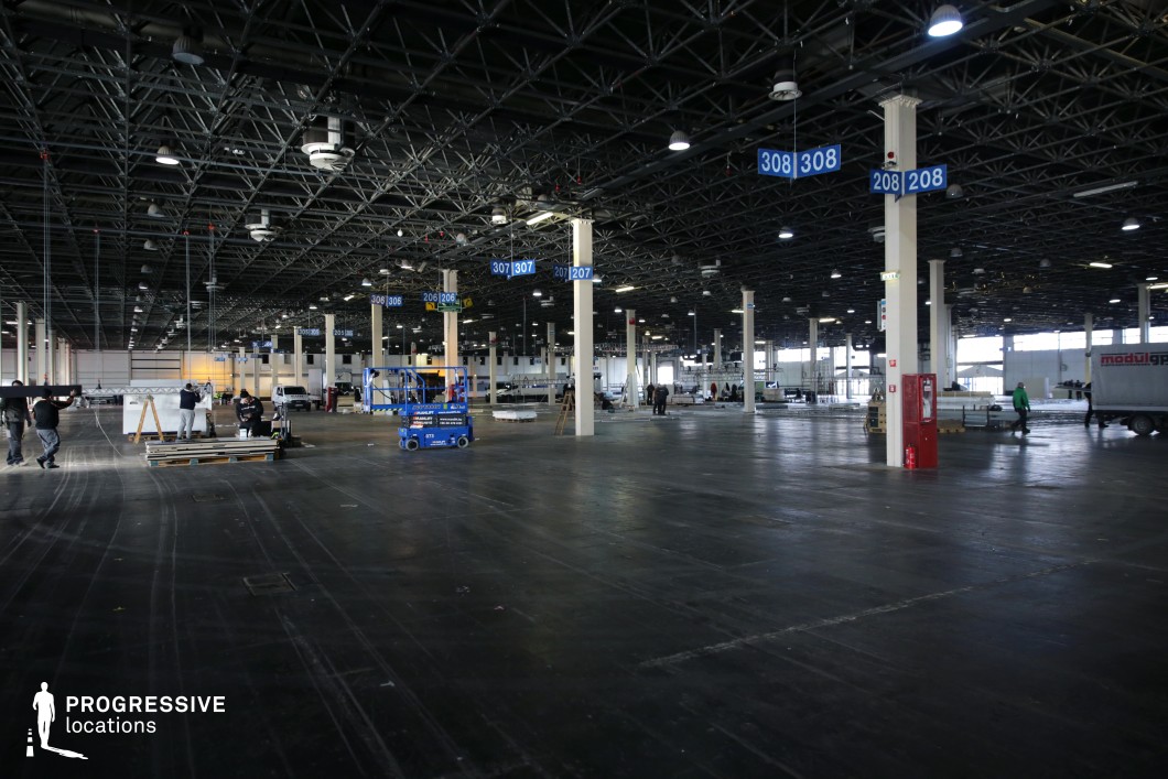 This immense industrial hall features a dark, grid-like ceiling with exposed metal beams and suspended lighting fixtures, creating a raw, utilitarian atmosphere. The open floor is made of dark concrete or heavy-duty material, marked with subtle traces of wear. White structural columns are evenly spaced throughout the space, with numbered blue signs providing a sense of order and segmentation. Large windows along the perimeter allow natural light to filter in, balancing the artificial lighting overhead. The vastness and modular design of this hall make it an ideal setting for dystopian films, large-scale event scenes, warehouse operations, or underground gatherings in action, thriller, or cyberpunk-inspired productions.