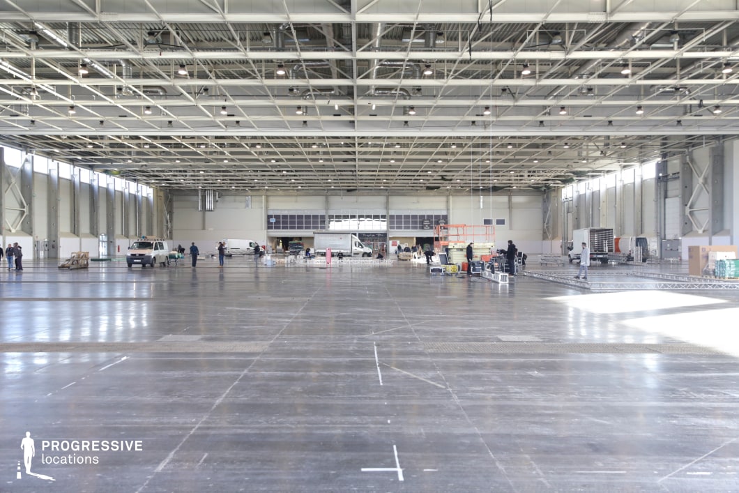 This massive industrial hall features an open, warehouse-style layout with a high metal ceiling supported by an intricate grid of beams and exposed ductwork. The polished concrete floor reflects the overhead lighting, emphasizing the vastness of the space. Large rolling doors and tall windows along the walls allow for natural light, contributing to the hall’s functional and utilitarian aesthetic. The location is ideal for action sequences, large-scale event settings, or dystopian and sci-fi productions. Its spacious and adaptable nature makes it perfect for staging logistical operations, underground gatherings, or high-stakes confrontations in a corporate or industrial environment.