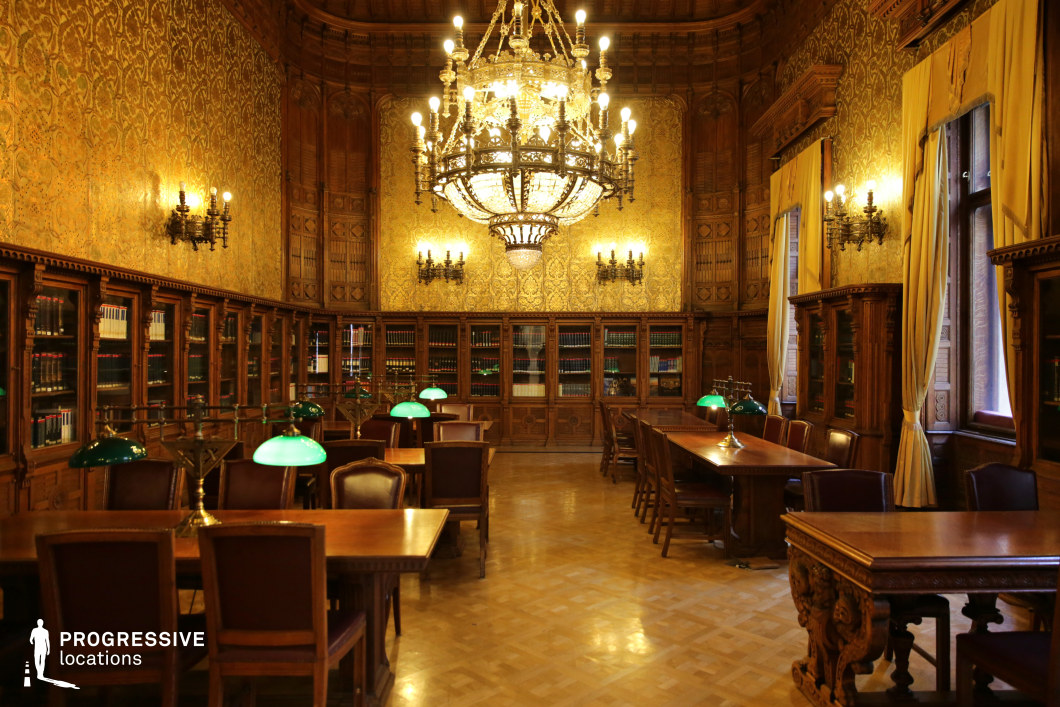 This richly decorated library exudes an air of sophistication, featuring dark wood paneling, intricately carved bookshelves, and golden damask-patterned wallpaper that enhances the luxurious aesthetic. A massive crystal chandelier dominates the space, casting a warm, ambient glow over the polished parquet flooring and the long wooden study tables equipped with classic green banker’s lamps. The high arched windows, draped with heavy curtains, allow for soft natural light to filter through, adding depth to the setting. The grandeur and historical ambiance make this location ideal for period dramas, intellectual or mystery-themed films, and scenes requiring an old-world, aristocratic library setting.