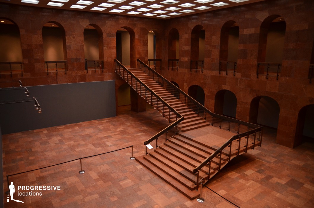 This striking atrium is defined by its rich red stone surfaces, giving it a bold yet austere aesthetic. The symmetrical design features a grand central staircase with sleek metal railings, flanked by arched openings on both levels, creating a structured and imposing atmosphere. The polished stone flooring and walls enhance the monochromatic palette, while the ceiling’s grid of recessed lighting provides uniform illumination. The space’s minimalistic yet powerful design makes it ideal for filming high-stakes drama, psychological thrillers, or corporate intrigue. It is particularly suited for scenes involving confrontations, power struggles, or enigmatic gallery settings.