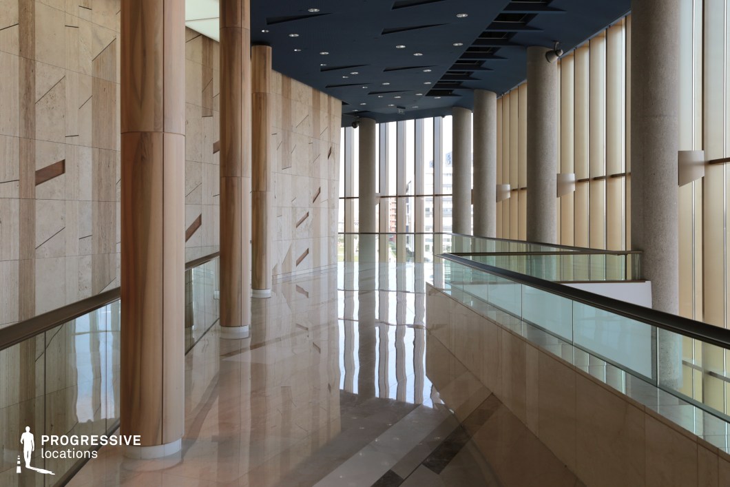 This sleek, modern atrium features a gently curving walkway lined with glass railings, polished marble flooring, and a series of grand wooden columns. The walls are adorned with light beige marble panels, intersected by subtle geometric inlays, adding depth and texture. A row of tall windows with vertical blinds allows soft, diffused light to enter, enhancing the sophisticated ambiance. The ceiling, painted in a deep blue hue, contrasts with the light-toned surfaces and incorporates recessed lighting. The combination of organic wood, smooth stone, and glass elements creates a refined, futuristic aesthetic. This setting is well-suited for corporate dramas, political thrillers, or high-end fashion shoots, providing a visually dynamic backdrop for professional, power-driven, or contemplative scenes.