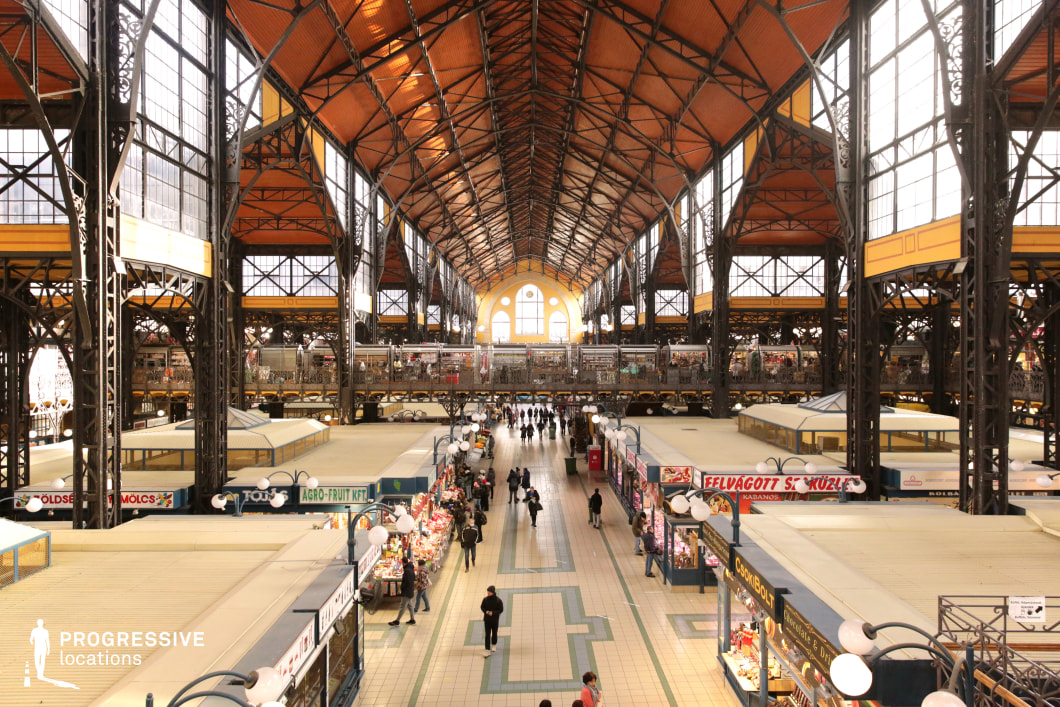 This expansive historic market hall features a striking wrought-iron framework supporting a high, arched ceiling with warm-toned wooden panels and large industrial-style windows that flood the space with natural light. The symmetrical design, intricate metal detailing, and elevated walkways create a visually dynamic setting, while the tiled floors and neatly arranged vendor stalls evoke a classic European market atmosphere. The market's multi-level structure offers a rich depth of field, making it an ideal location for filming scenes set in bustling urban environments, period dramas, or noir-inspired thrillers. Its grand architectural scale and timeless aesthetic lend themselves well to depictions of trade, commerce, or clandestine meetings in a vibrant, historic setting.