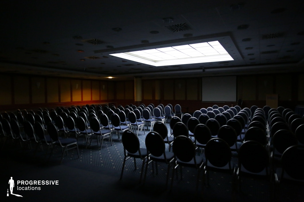 This traditional conference hall features rows of dark blue cushioned chairs arranged in a symmetrical theater-style setup, facing a projector screen and podium. The space is enclosed by warm wooden paneling and a low, grid-patterned ceiling with recessed lighting. A large, centrally located rectangular skylight provides an eerie, uneven illumination, casting shadows throughout the room. The deep blue carpet enhances the moody atmosphere, making this location ideal for tense, suspenseful, or mystery-driven film scenes. It is well-suited for corporate dramas, conspiracy thrillers, or unsettling psychological narratives, offering a setting that can transition from formal to ominous depending on the lighting and tone.